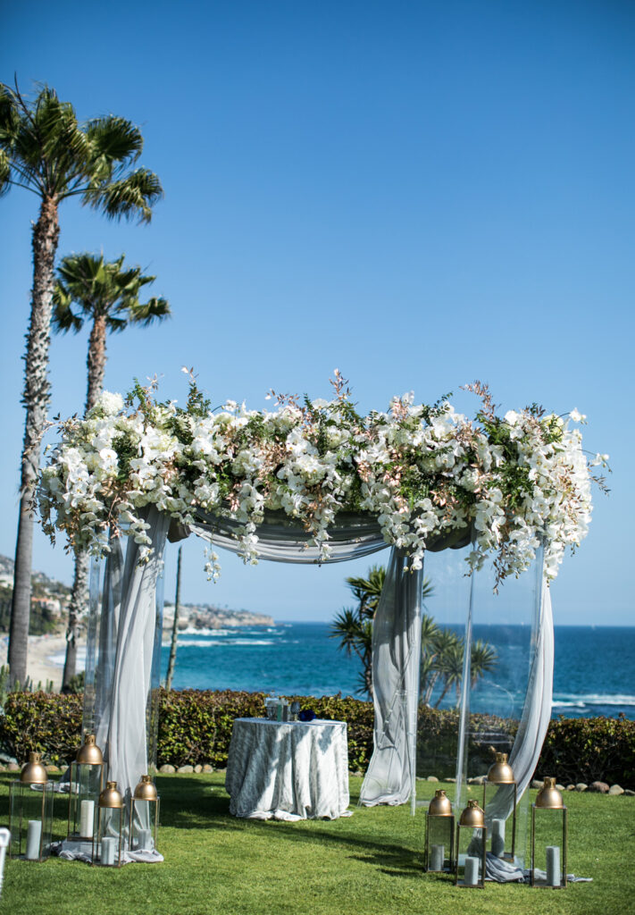 Luxurious seaside wedding arch decorated with lush white and pink florals and flowing drapes, complemented by elegant lanterns and a table with blue glassware, set against a backdrop of palm trees and the ocean.