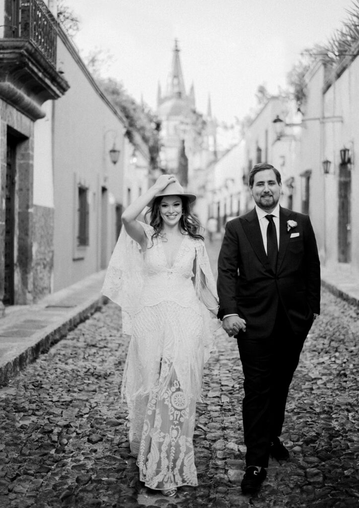 Black and white photo of a joyful bride in a lace dress and a groom in a suit, walking hand in hand on a cobblestone street in San Miguel, with historic buildings and a spired church in the background.