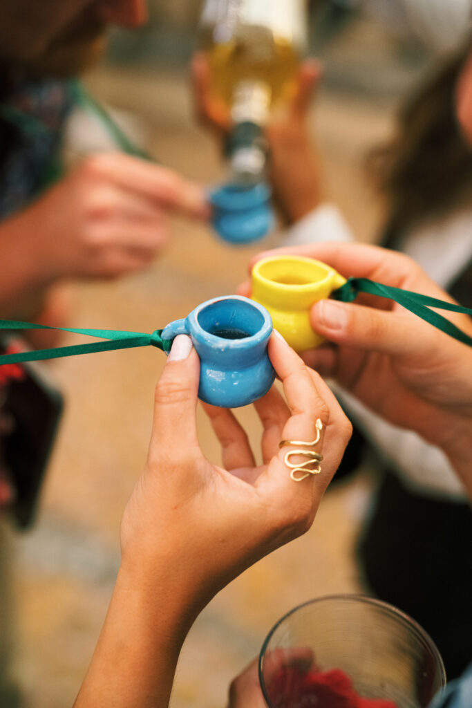 Close-up of hands holding colorful ceramic tequila cups attached to a green cord, during a festive tequila tasting event, with a bottle of tequila being poured in the background.
