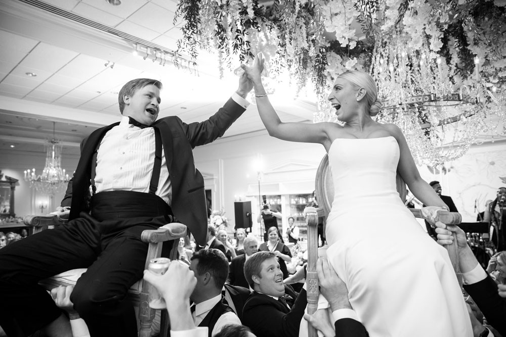 Black and white image of the joyous bride and groom being lifted on chairs during the wedding reception, surrounded by cheering guests.