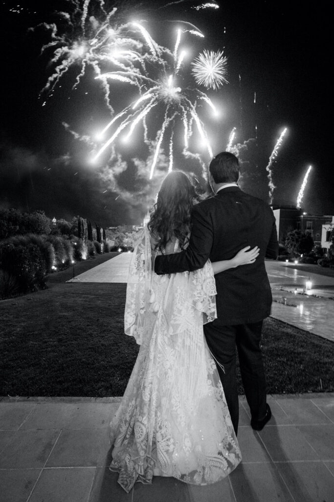 Black and white photo of a bride and groom from behind, embracing and watching a spectacular fireworks display in the night sky, celebrating their wedding day.