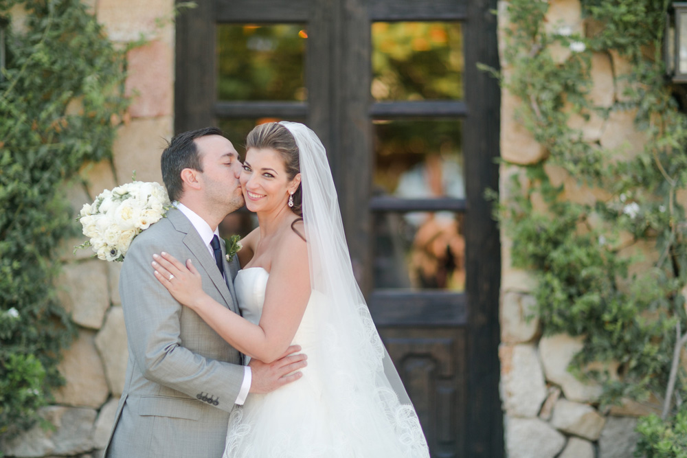 Groom in a light gray suit affectionately kissing the bride on the cheek, who is smiling and holding a bouquet of white flowers, in front of a rustic stone wall with ivy and a dark wooden door.