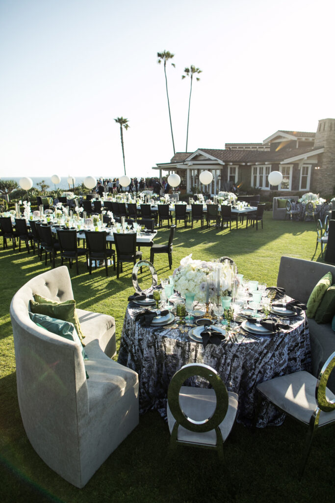 Outdoor wedding reception on a manicured lawn with elegant table settings featuring dark chairs, floral centerpieces, and sophisticated tableware, underlined by tall palm trees with a building in the background, bathed in soft sunset light.