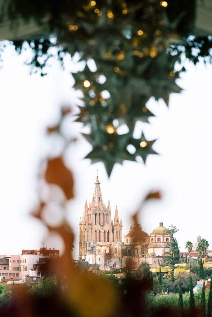 Artistic view through blurred foreground leaves, focusing on the intricate spires of a historic church in San Miguel, framed by twinkling lights, against a backdrop of colorful buildings.
