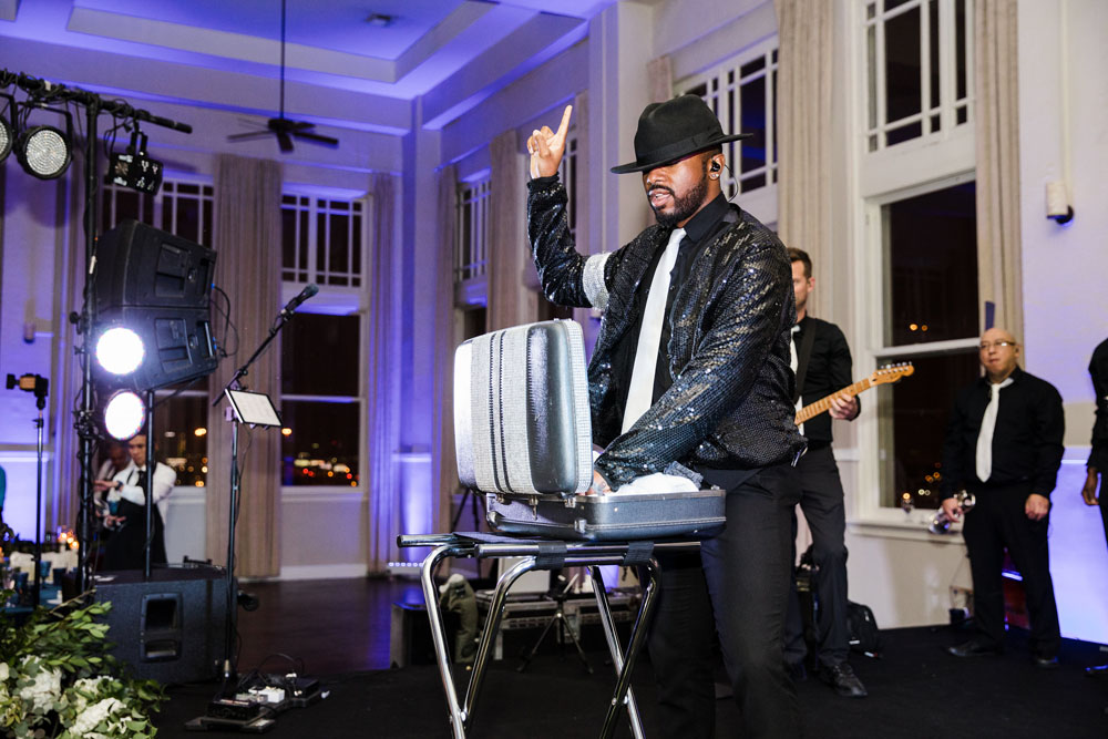 Charismatic DJ in a black sequined jacket hyping up the wedding guests, with a live band playing in the softly lit background.