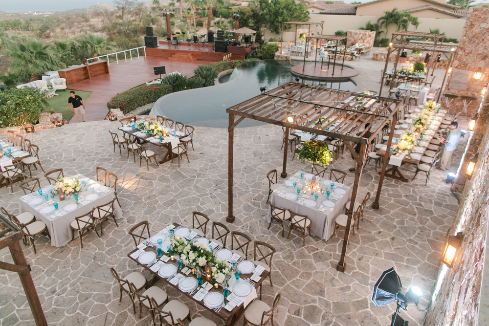 Elevated view of a wedding reception area in Cabo, showcasing elegantly set tables on a stone floor, an infinity pool, and a wooden deck patio area, arranged for a luxurious evening event, awaiting guests.