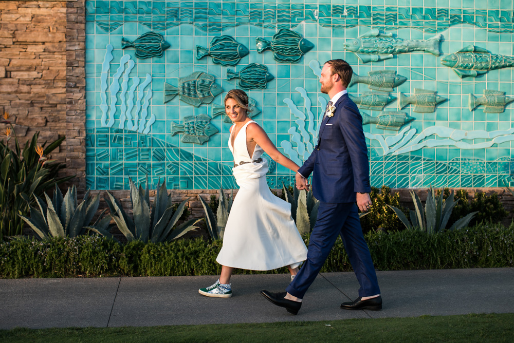 Bride in a stylish white dress with sneakers and groom in a navy suit walking hand-in-hand in front of a turquoise mural depicting fish.