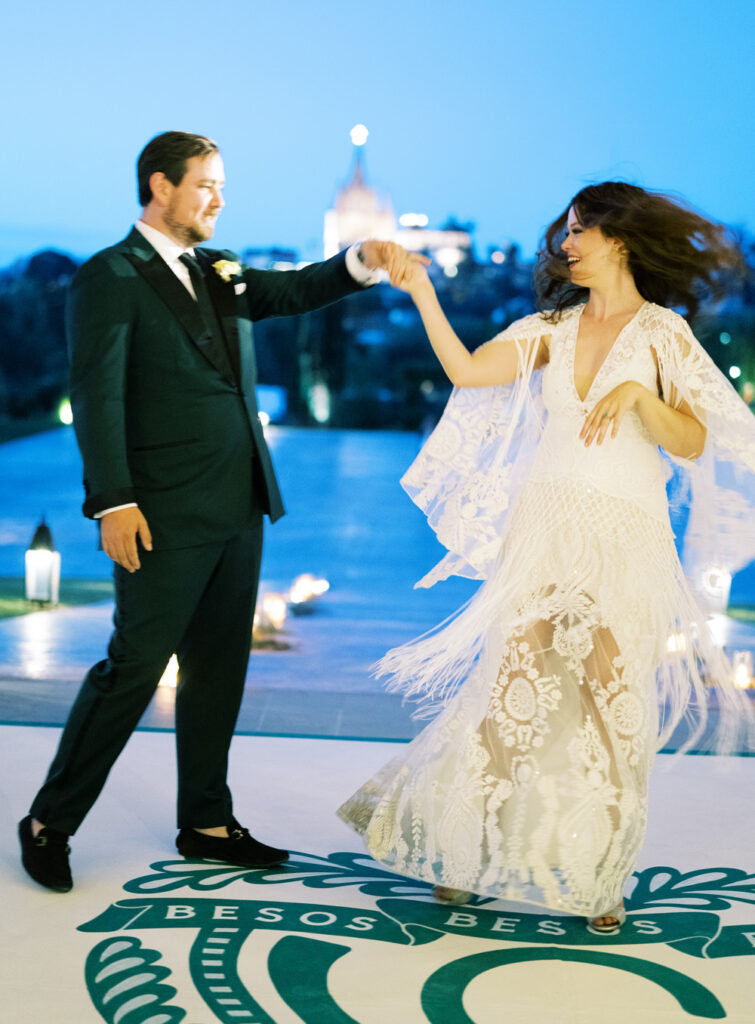 Bride in a detailed lace dress and groom in a dark suit dancing joyfully on a dance floor with 'BESOS BESOS' design, with a softly illuminated church in the background at dusk.