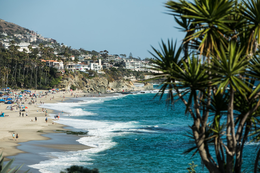 Scenic view of a bustling beach in Orange County, California, featuring waves crashing onto the sandy shore, beachgoers enjoying the sunny day, and residential houses nestled among lush greenery and palm trees on the hillside.