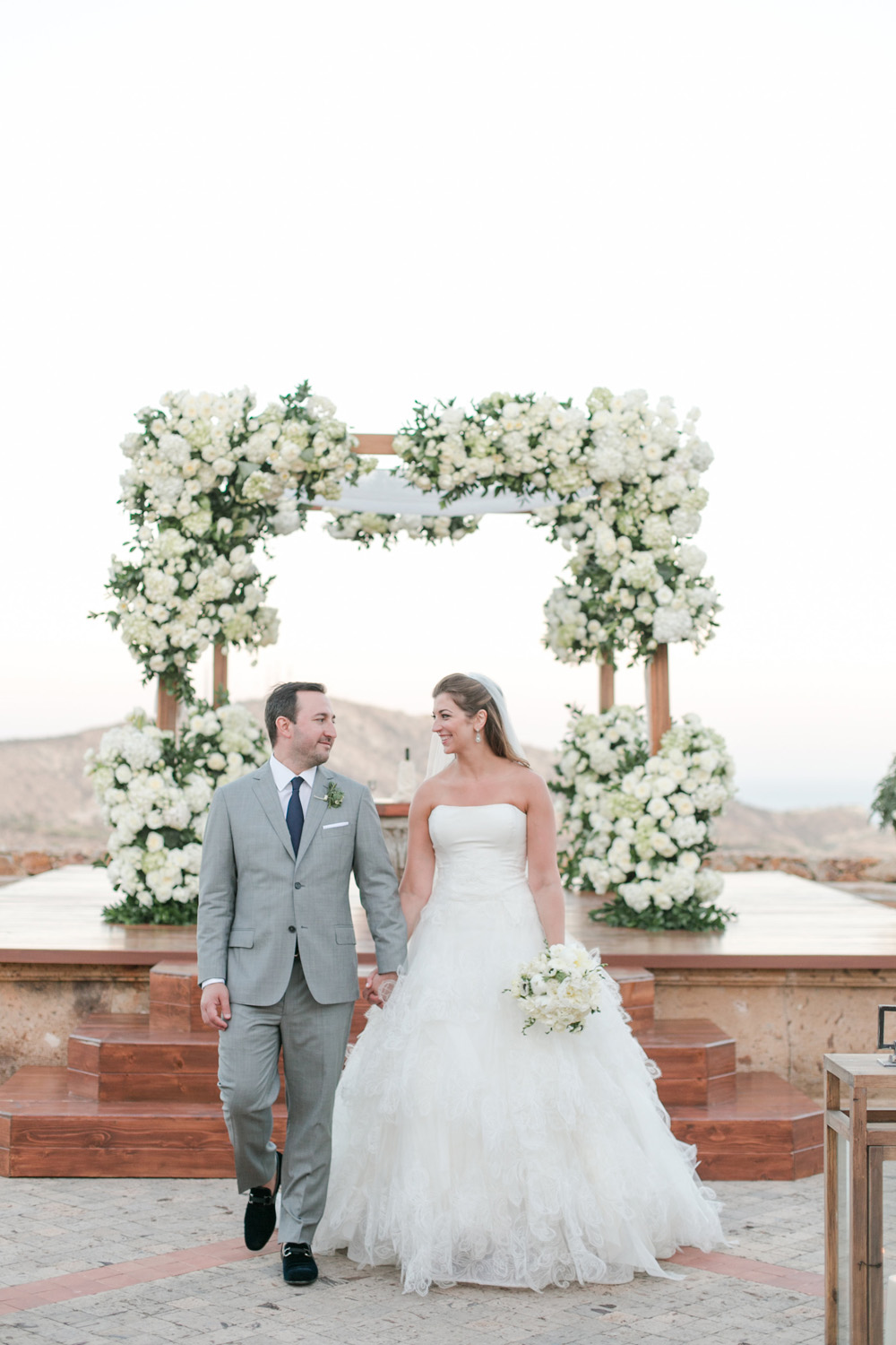 Bride in a flowing white gown and groom in a light gray suit holding hands and smiling at each other as they walk away from an altar adorned with lush white floral arrangements, set against a scenic outdoor backdrop.