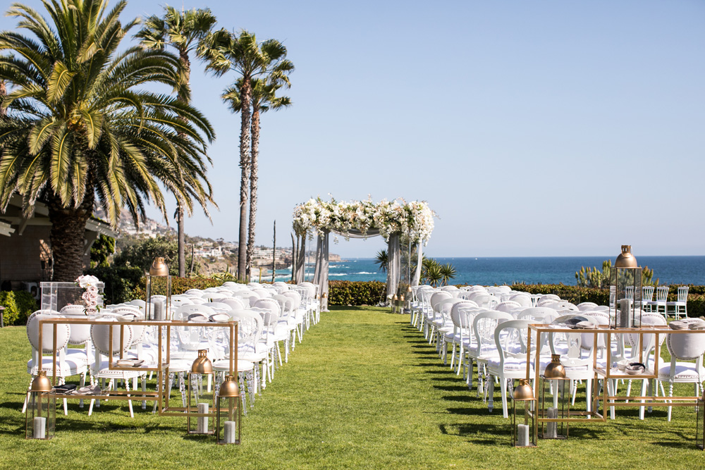 Idyllic wedding ceremony setup on a lush lawn overlooking the ocean, featuring rows of white chairs, a floral arch at the altar, and tall palm trees swaying against a clear blue sky at Pelican Hill.