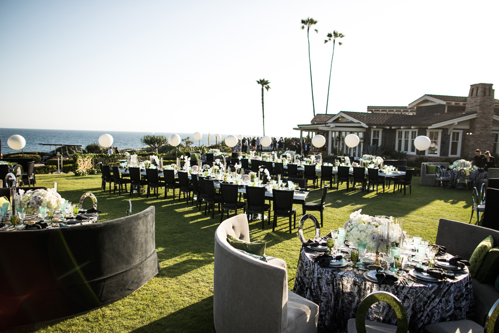 Outdoor wedding reception setup at Montage Laguna Beach on a lush lawn overlooking the ocean, featuring black tables with luxurious floral centerpieces, elegant dark chairs, and plush lounge areas, all under the soft glow of sunset light with palm trees in the background.