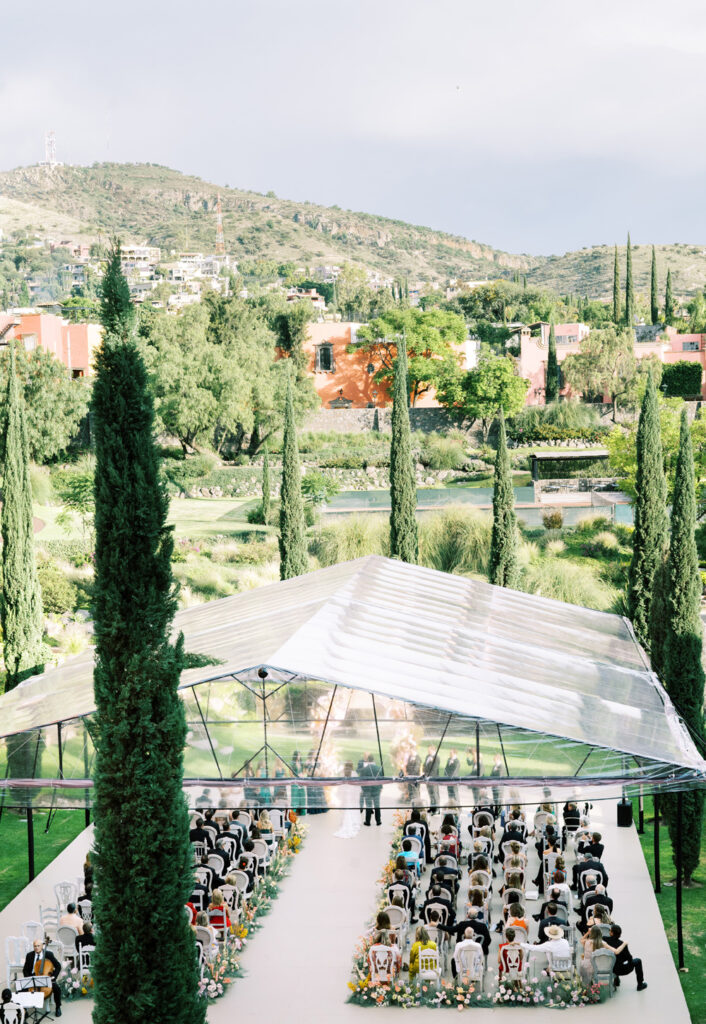 Elevated view of a wedding ceremony at Rosewood Hotel, featuring guests seated in chairs, under a transparent tent, surrounded by scattered cypress trees and a lush green hillside background.