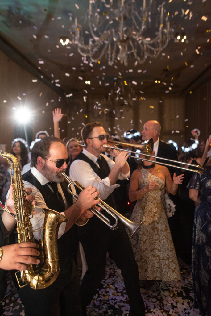 Excited band with brass instruments engaging with guests on the dance floor, under the grand chandelier of the wedding venue.