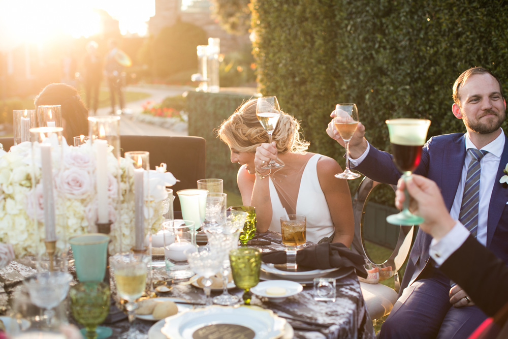 Bride and groom toasting at their wedding reception, sitting at a table adorned with a floral centerpiece and elegant tableware, bathed in the golden glow of sunset.