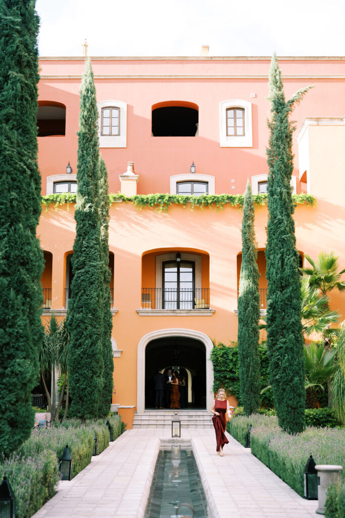 Symmetrical view of the Rosewood Hotel in San Miguel, showcasing a vibrant salmon-colored building framed by tall cypress trees and adorned with hanging green ivy, with a reflective water feature leading to an arched doorway, and a woman in a red dress walking beside it.