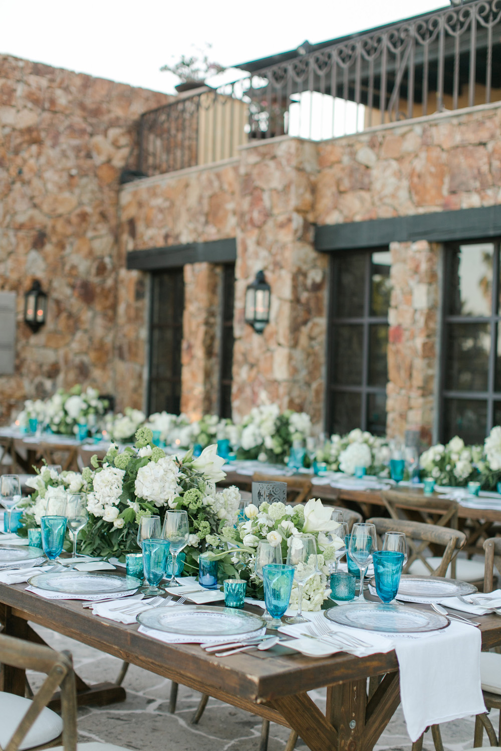 Elegant wedding reception setup featuring rustic wooden tables adorned with white floral centerpieces and blue transparent glasses, set against the backdrop of a stone building with dark framed windows.