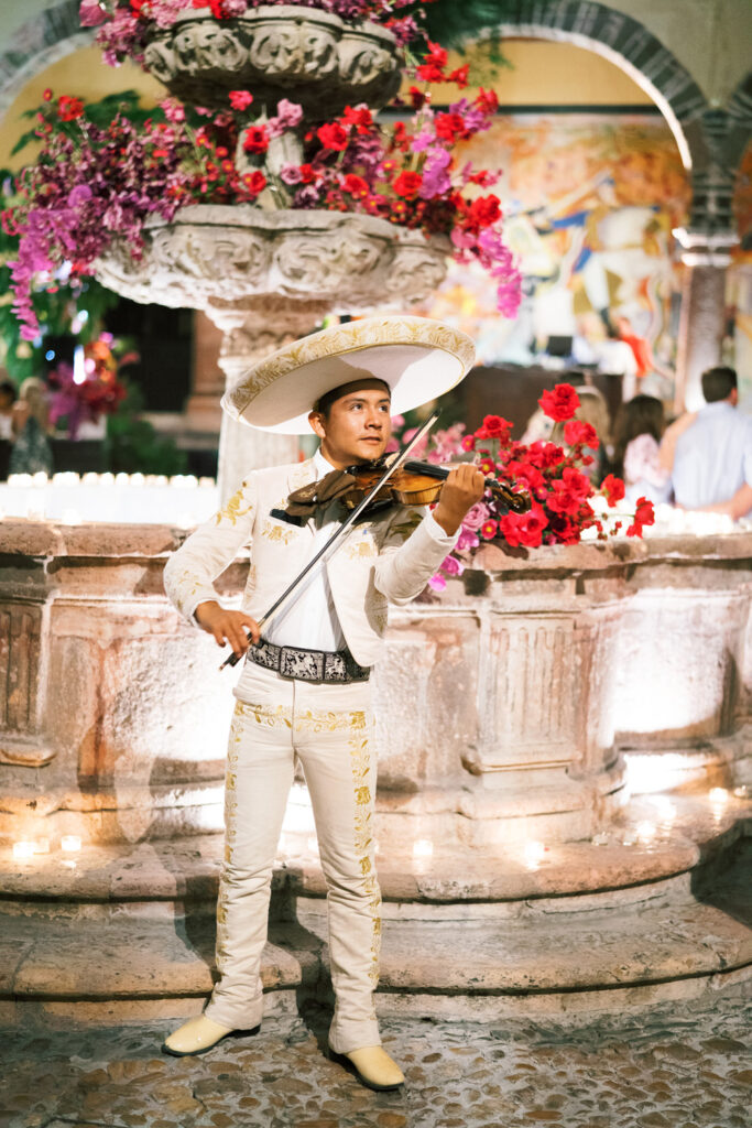 Mexican mariachi violinist in traditional white embroidered suit and sombrero, playing energetically at a wedding with a vibrant floral fountain and mural backdrop, adding a festive ambiance to the celebration.