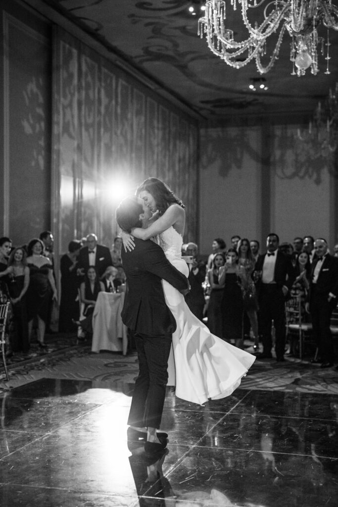 Black and white photograph of the bride and groom sharing a romantic kiss on the dance floor, with guests and elegant chandeliers in the background.