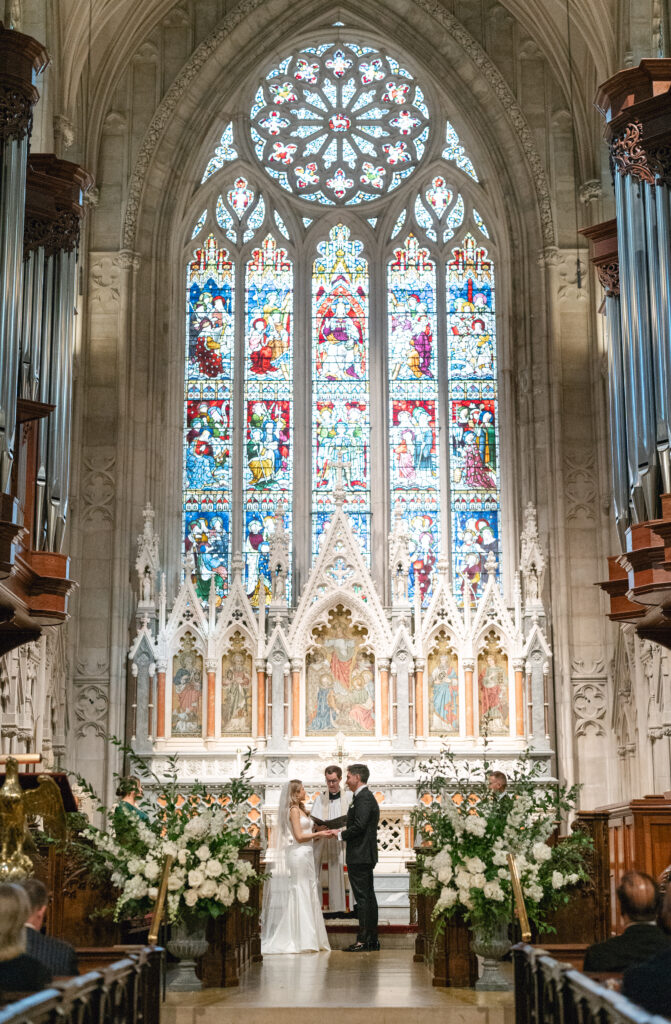 Bride and groom standing at the alter inside Grace Church in NYC, with a large stained glass window behind them