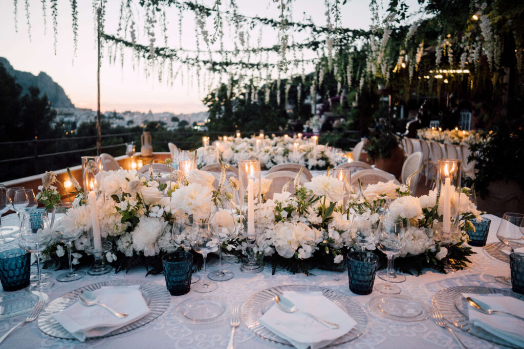 Table covered in tapered candles, white flowers, and blue glassware. Overlooking the town of Capri, Italy