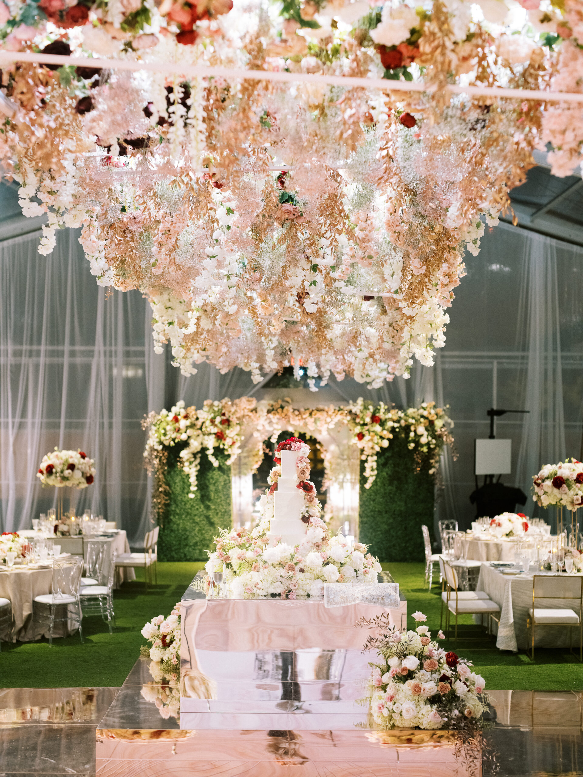 Large floral arrangement hunt from the ceiling of a clear tent, hanging over a white wedding cake covered in roses