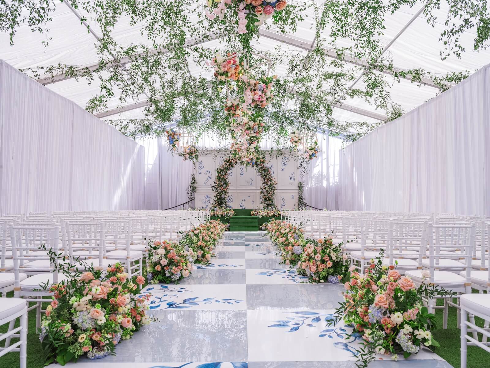 A wedding ceremony inside a tent, with greenery and flowers hanging from the ceiling, flowers lining the aisle, and a checkered gray and white floral floor at Rosewood Mansion in Dallas