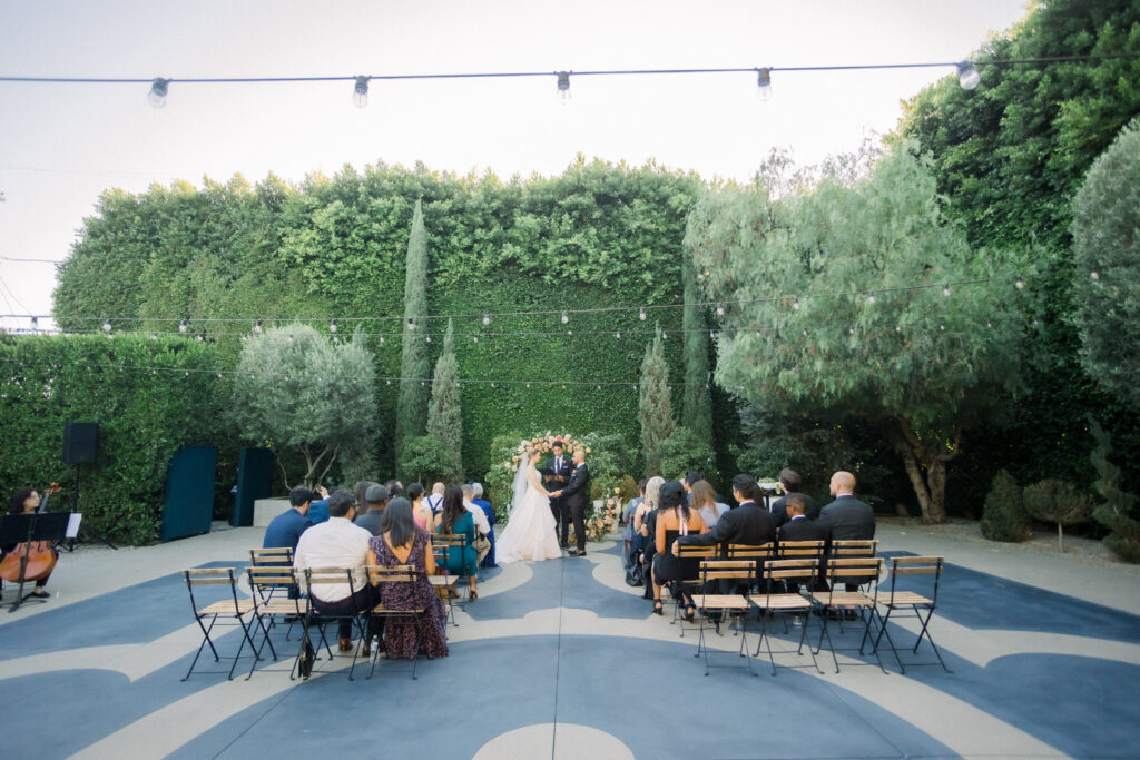 Bride and groom standing in front of large green hedges, under cafe lights, outdoors at their wedding ceremony at The Fig House